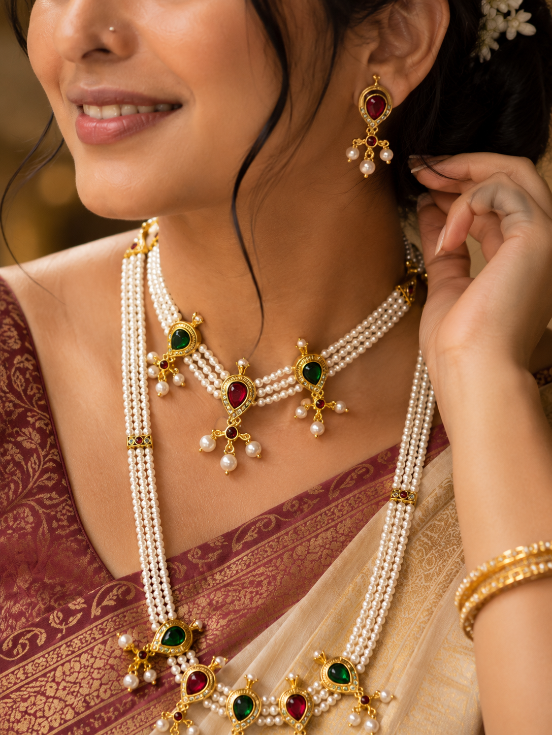 A close-up lifestyle shot of a woman wearing an elaborate traditional Indian gold-plated jewelry set with white pearls and green, red, and gold accents. The set includes a multi-strand necklace, matching drop earrings, and a gold bangle, styled with a maroon and beige saree.
