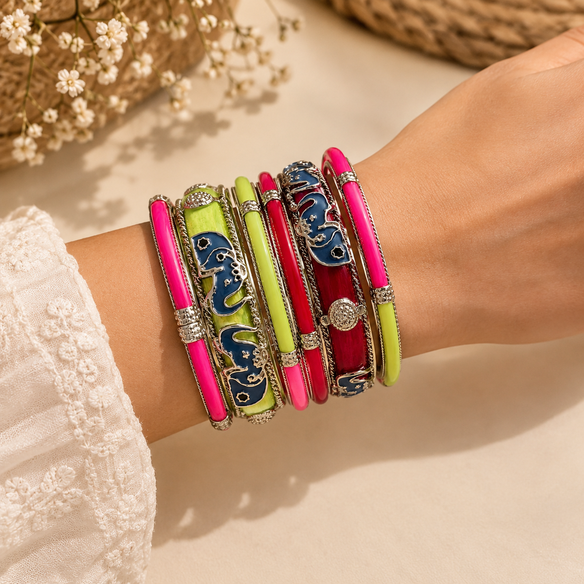A close-up of a person's wrist wearing a stack of ornate bangles. The bracelets feature bright pink, lime green, and red enamel panels with intricate blue and silver patterns, along with silver-toned metal borders and rhinestone accents.