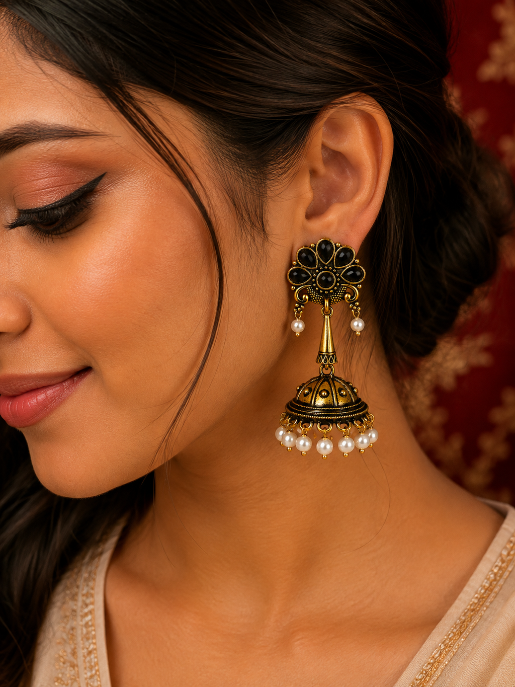A close-up of a woman wearing a traditional Indian gold and black jhumka earring with white pearl accents, set against a warm red patterned background.