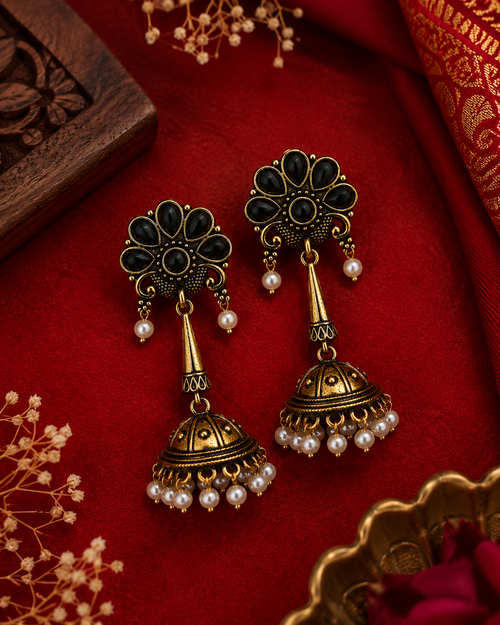 A pair of ornate gold-toned jhumka earrings with black stone accents and small pearl beads, displayed on a rich red fabric background with decorative floral and wooden props.