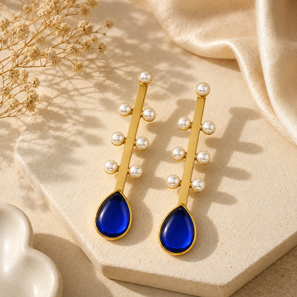 A pair of gold drop earrings with small white pearl beads and a deep blue teardrop stone, displayed on a beige stone slab with draped satin fabric and dried flowers.
