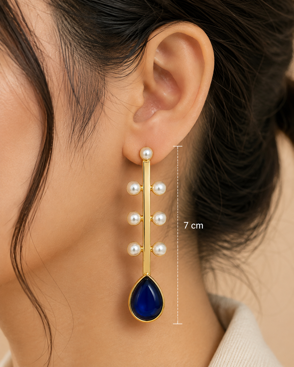 Close-up of a model's ear wearing a gold dangle earring with white pearls and a deep blue teardrop stone. A vertical measurement marker indicates the earring length is 7 cm.
