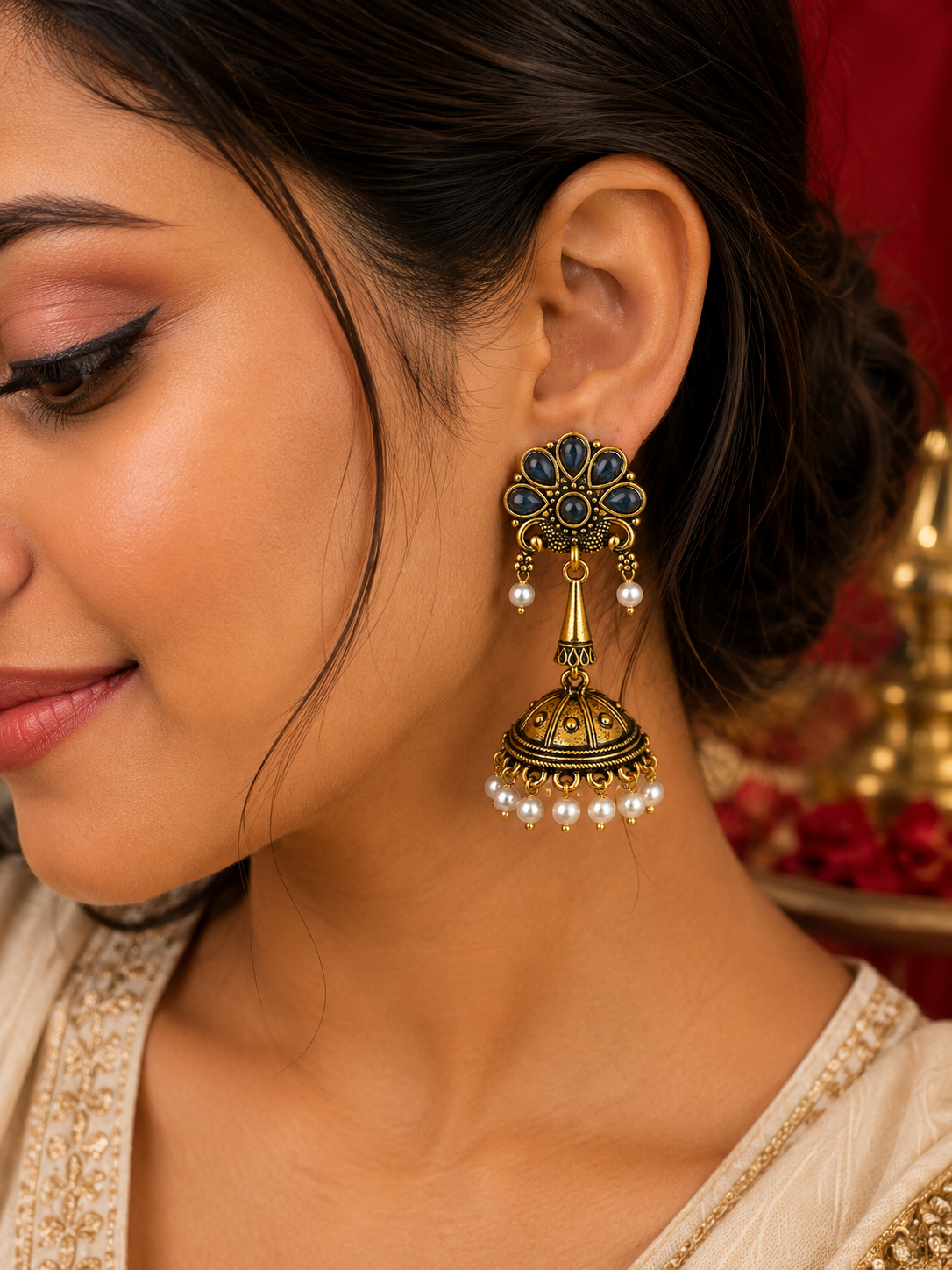 A close-up of a woman wearing a traditional Indian gold-plated jhumka earring with dark blue enamel, gold filigree, and dangling white pearl beads.