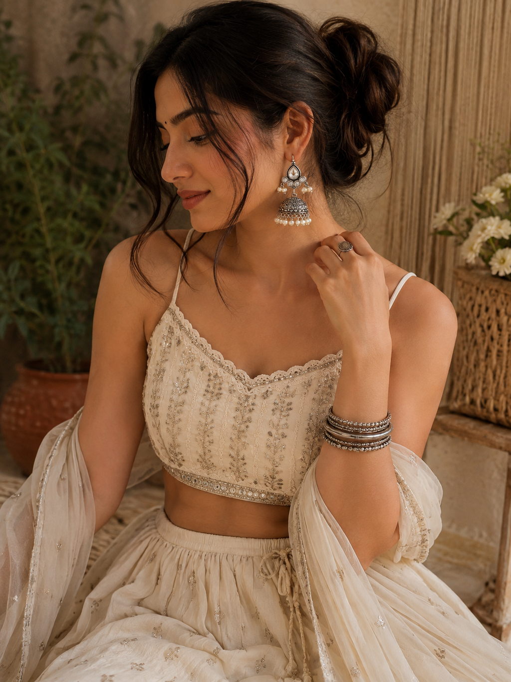 A woman wearing a cream-colored embroidered traditional outfit with a crop top and flared skirt, styled with matching sheer dupatta and ornate silver-toned earrings.