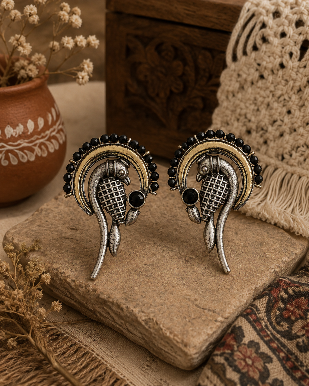 A pair of ornate silver and gold earrings with black bead accents and small black stones, displayed on a stone block with dried flowers and a textured textile in the background.