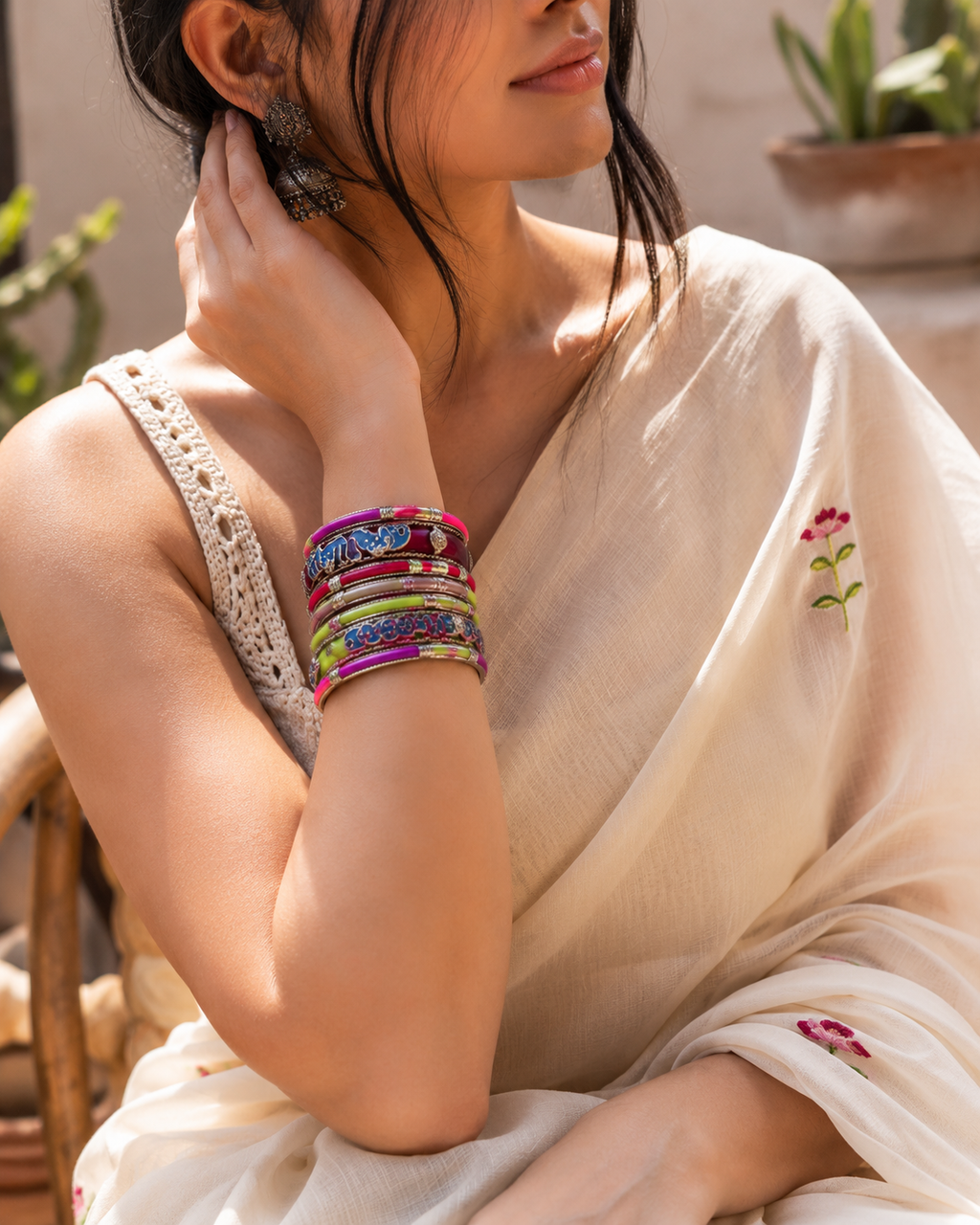 A woman wearing a cream-colored embroidered saree and a stack of colorful metallic bangles on her wrist, posed in a warm, sunlit indoor setting.