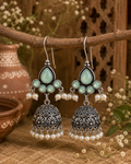 A pair of traditional Indian silver-toned jhumka earrings with mint green teardrop stones, small pearl bead clusters, and ornate filigree bell-shaped drops, hanging from a rustic wooden branch.