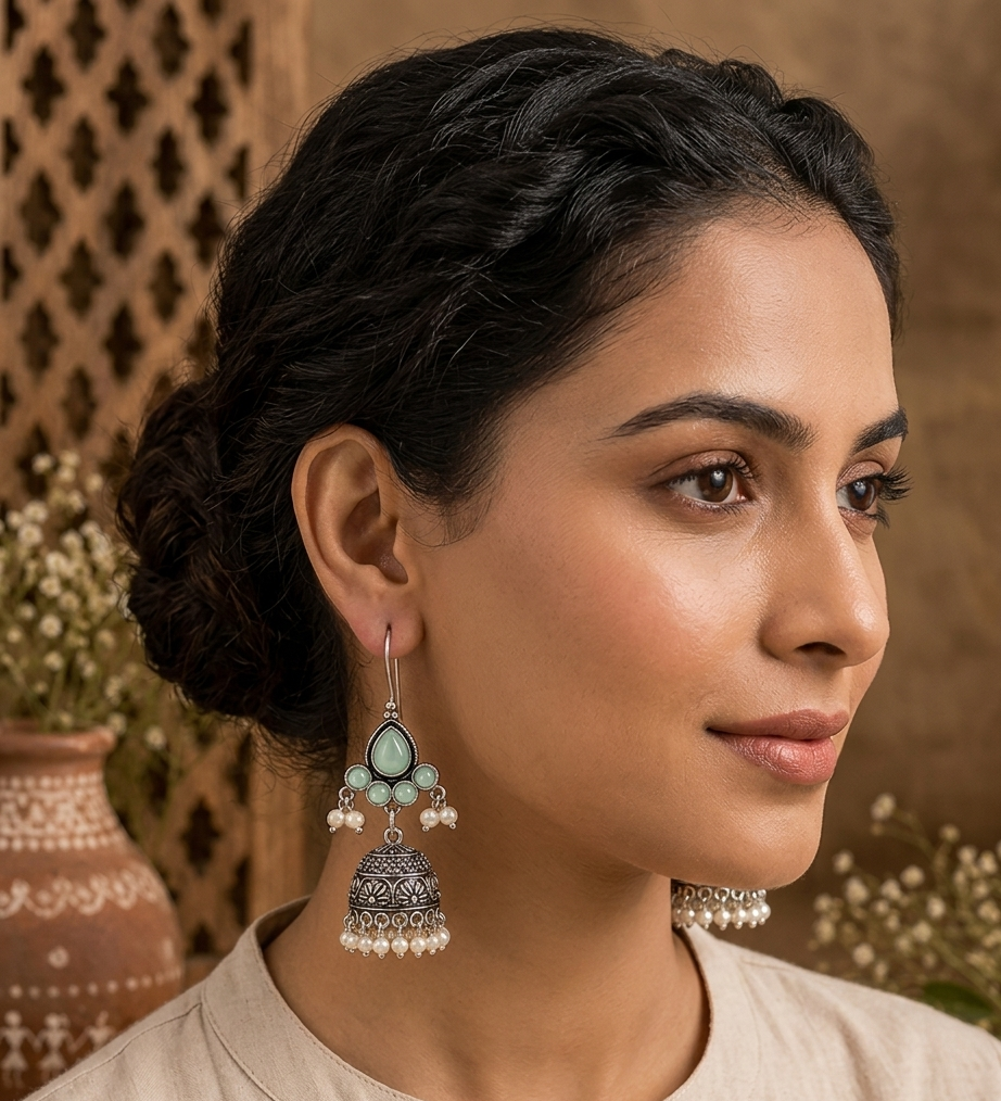 A woman wearing a pair of ornate silver-toned jhumka earrings with mint green stones and white pearl-like beads, shown in a warm, styled portrait setting.