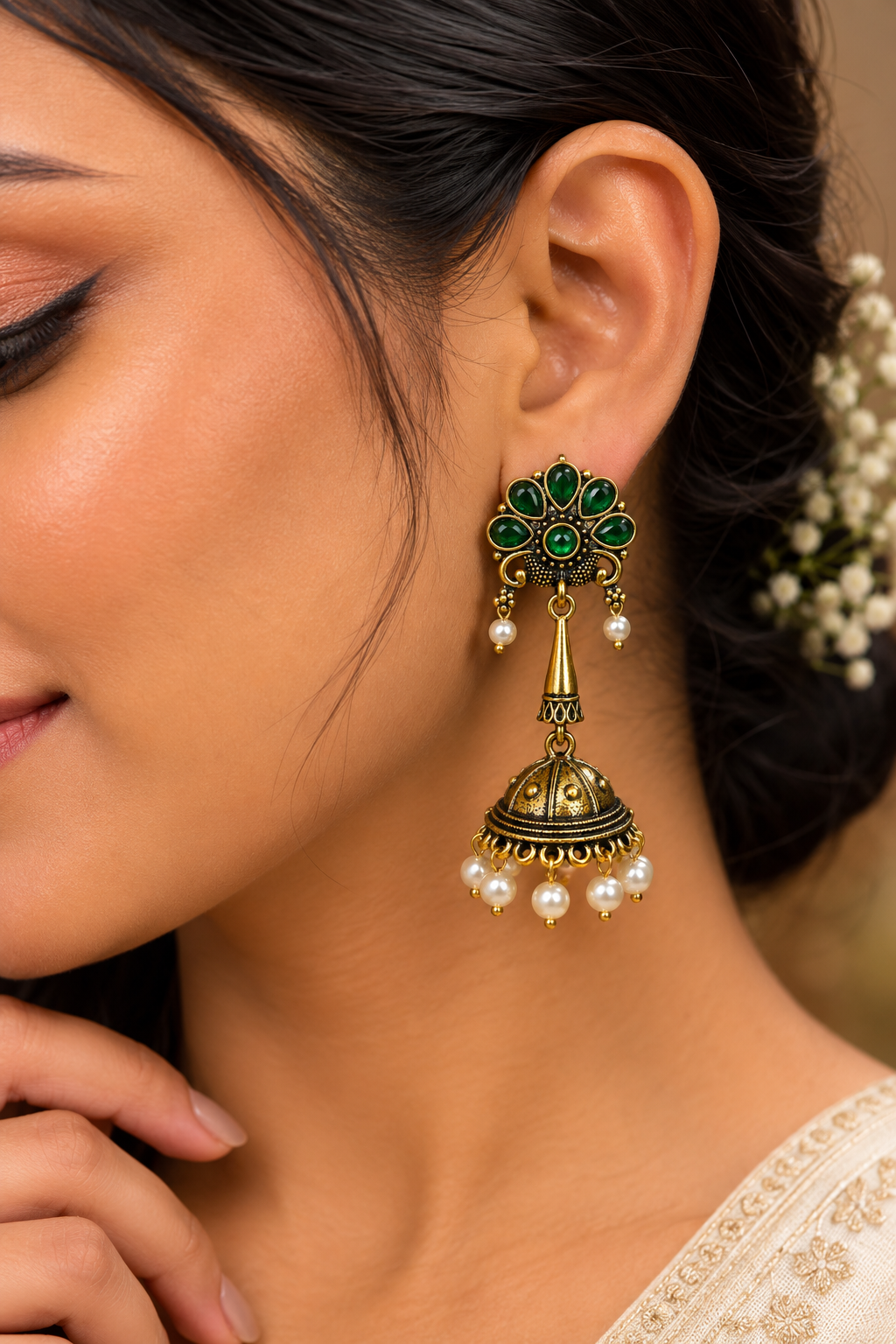 A close-up of a woman wearing an ornate gold and green jhumka earring with dangling pearl beads, shown on her ear against a softly blurred background.