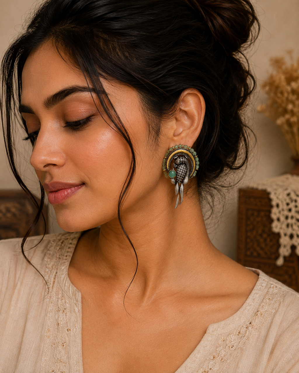 A close-up of a woman wearing a large, ornate silver-toned huggie hoop earring with green and gold accents and feather details, paired with a beige embroidered top.