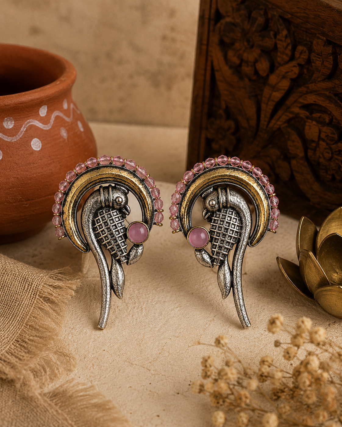 A pair of ornate Indian-style earrings with silver and gold-plated metalwork, pink stone accents, and a curved design, displayed on a textured beige surface with traditional props.