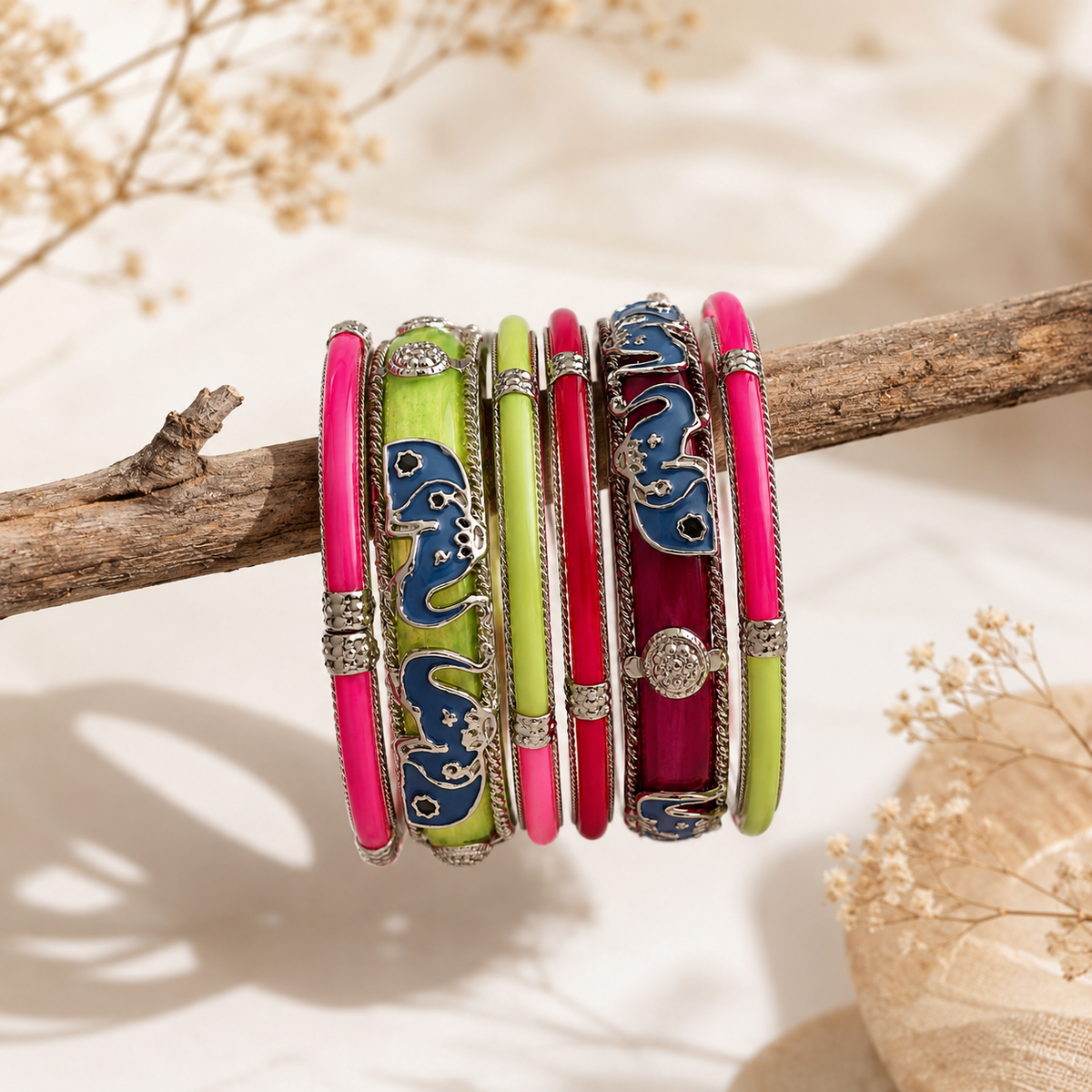 A set of six ornate bangles displayed on a rustic wooden branch. The bangles feature vibrant neon pink, lime green, and deep magenta enamel panels with detailed silver-toned metalwork, including elephant motifs and circular decorative elements.