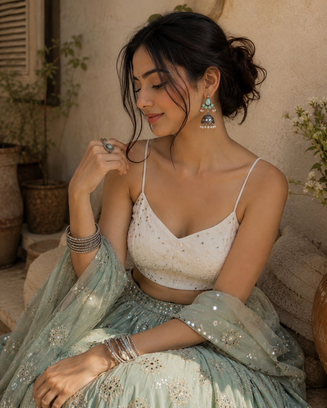 A woman wearing a light sage green and white embellished traditional outfit with matching sheer dupatta, seated in a warm, rustic setting.