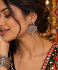 A close-up lifestyle shot of a woman wearing a large silver-toned jhumka earring with dangling beadwork and a small hanging charm, paired with a heavily embellished black and red traditional top.