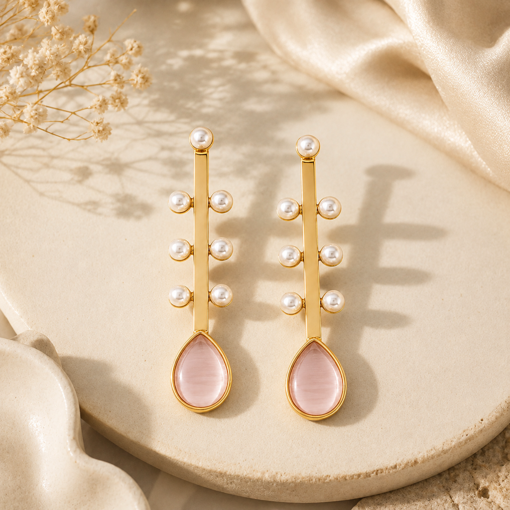 A pair of gold drop earrings with small white pearl beads and teardrop-shaped pink stone drops, displayed on a beige ceramic dish with soft satin fabric and dried flowers.