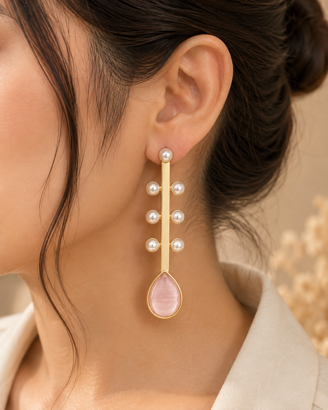 A close-up of a model wearing a long gold dangle earring with white pearls and a teardrop-shaped pink stone.