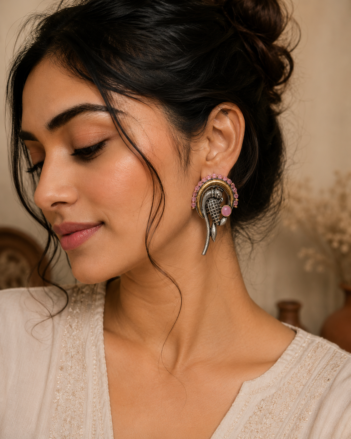 A close-up of a woman wearing a large, ornate silver-toned earring with pink gemstone accents and a small gold-toned circular detail, paired with a cream-colored embroidered top.