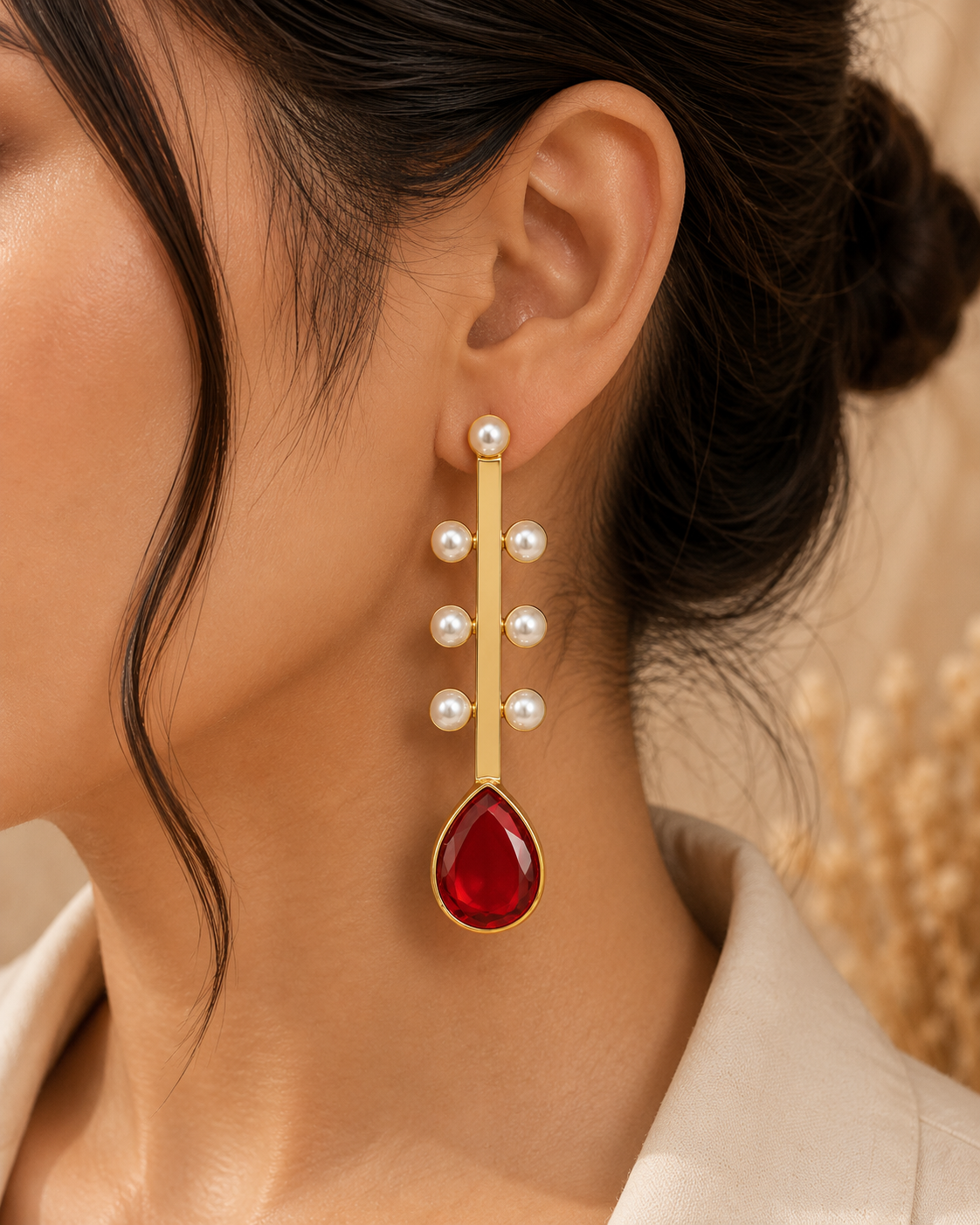 A close-up of a model wearing a long, elegant dangle earring. The earring features a vertical gold bar adorned with five white pearl-like beads, culminating in a large, teardrop-shaped red gemstone at the bottom, all set in a polished gold-tone metal.