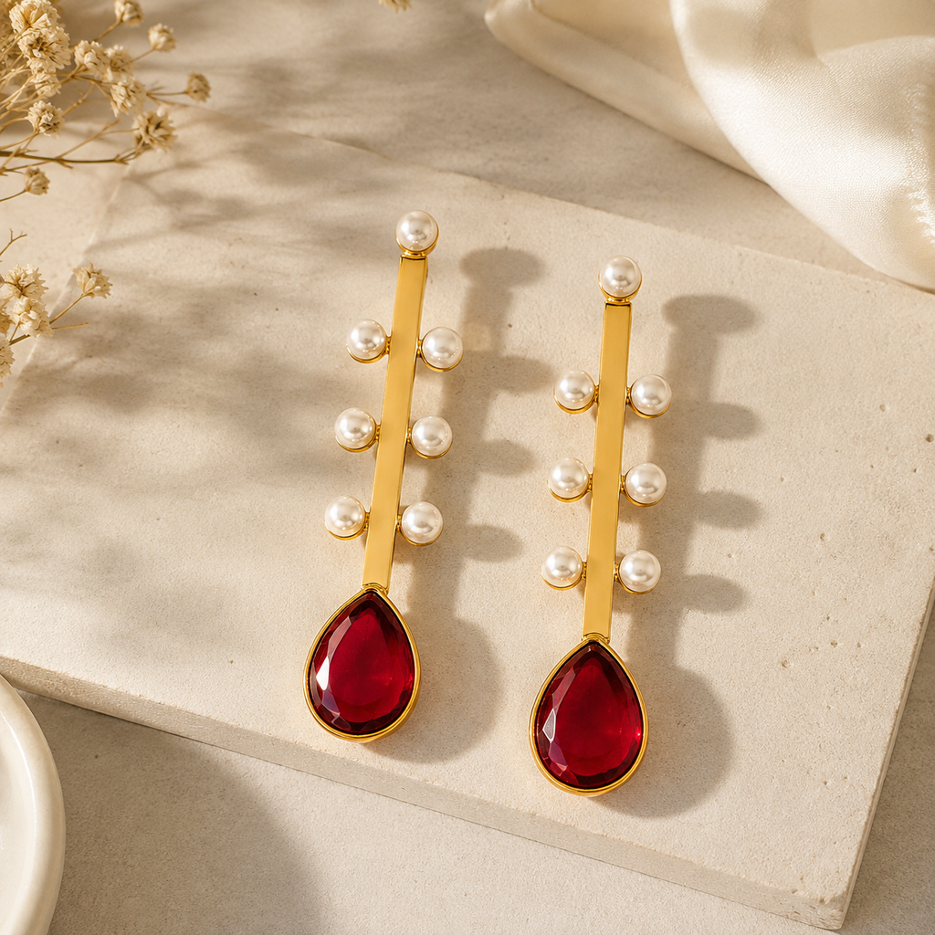 A pair of gold drop earrings with small white pearl beads and large teardrop-shaped deep red gemstones, displayed on a textured beige stone surface with soft fabric and dried flowers in the background.