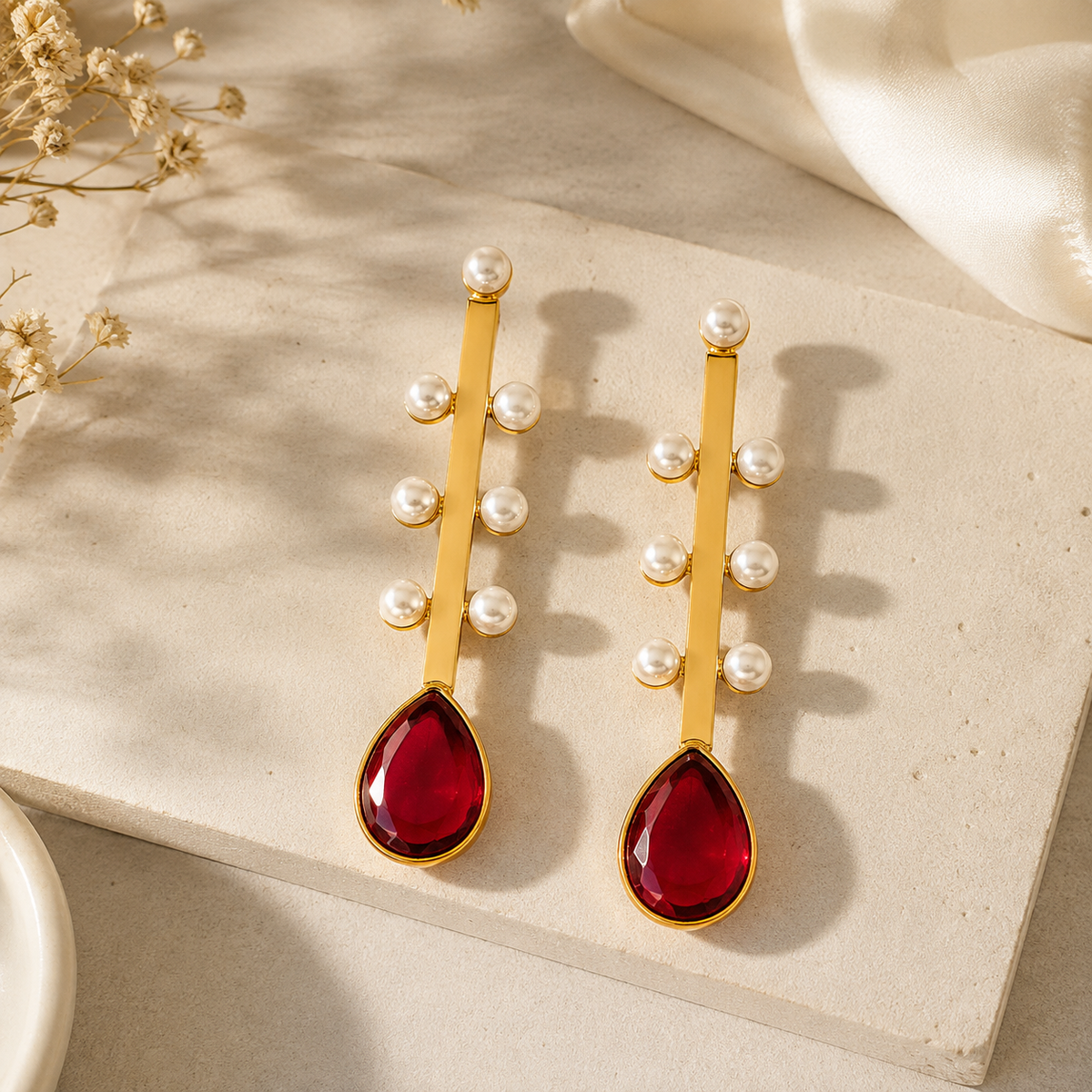 A pair of gold drop earrings with small white pearl beads and large teardrop-shaped deep red gemstones, displayed on a textured beige stone surface with soft fabric and dried flowers in the background.