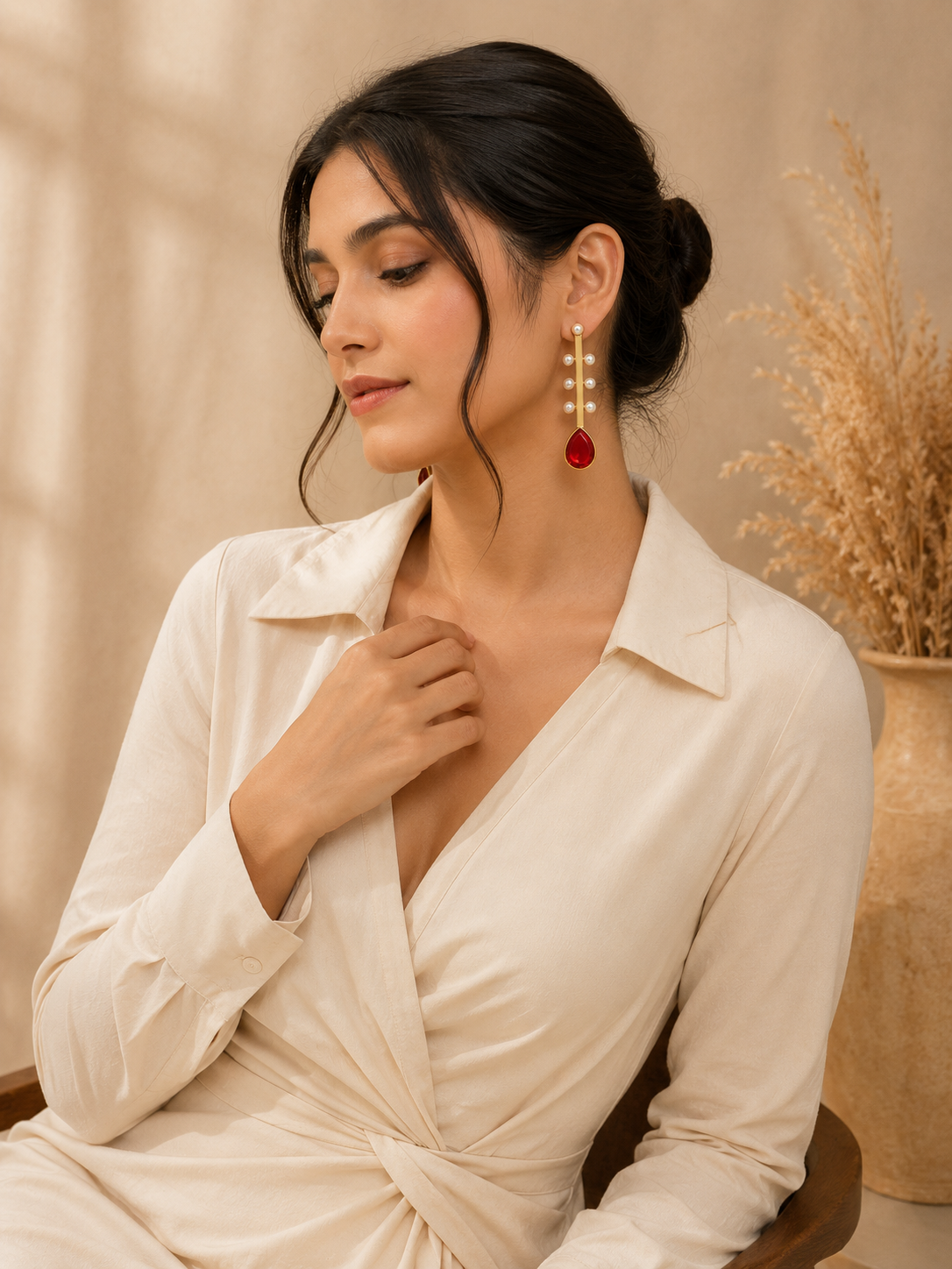 A woman wearing a cream-colored wrap dress and a gold drop earring with a red teardrop gemstone, posed in a warm, neutral-toned indoor setting.