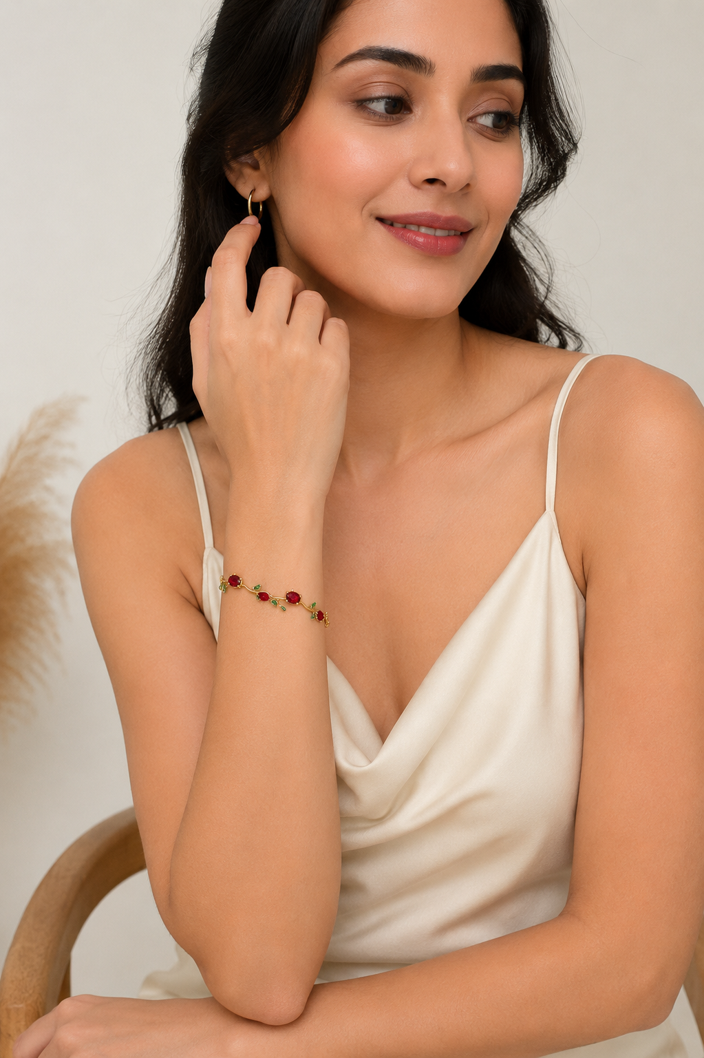 A woman wearing a delicate gold chain bracelet with small red and green bead accents, styled with a cream camisole top against a soft beige background.