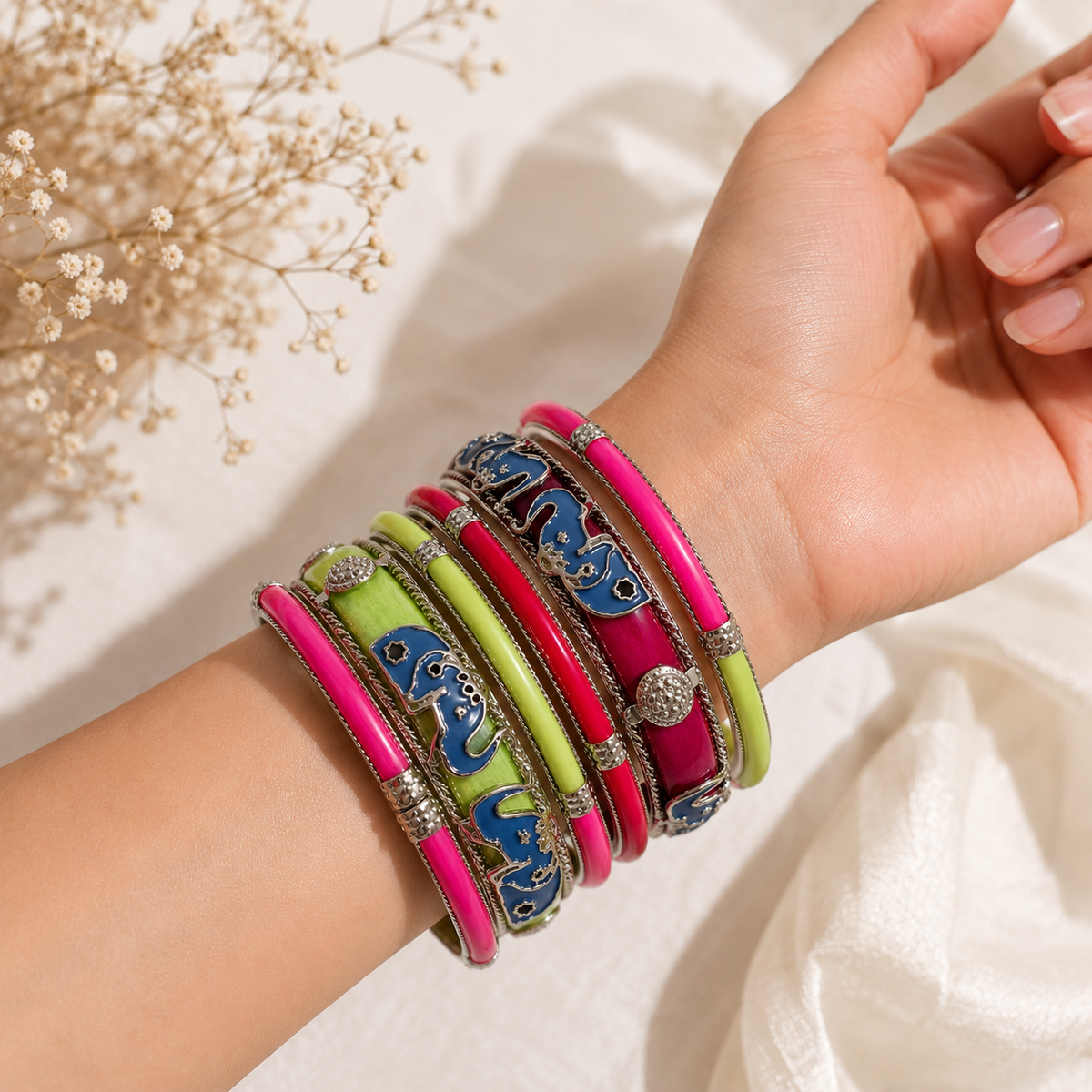 A close-up of a person's wrist wearing a stack of colorful bangles, including bright pink, lime green, magenta, and blue with silver-toned borders, against a soft white fabric background with dried flowers.