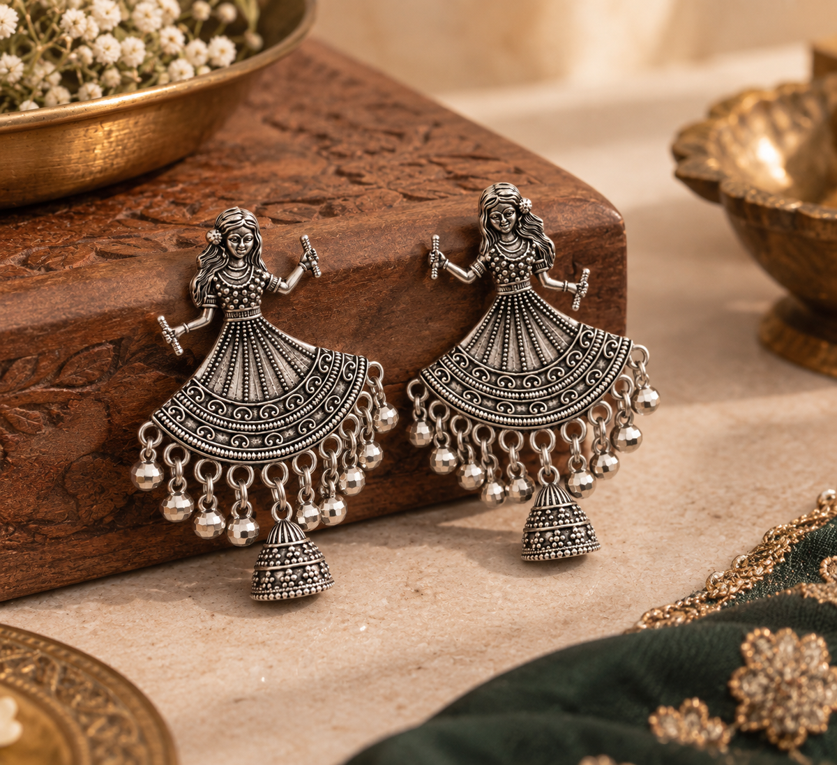 A pair of silver-toned traditional Indian dangle earrings shaped like dancing figures with ornate filigree details and dangling bell charms, displayed on a carved wooden block with decorative props.