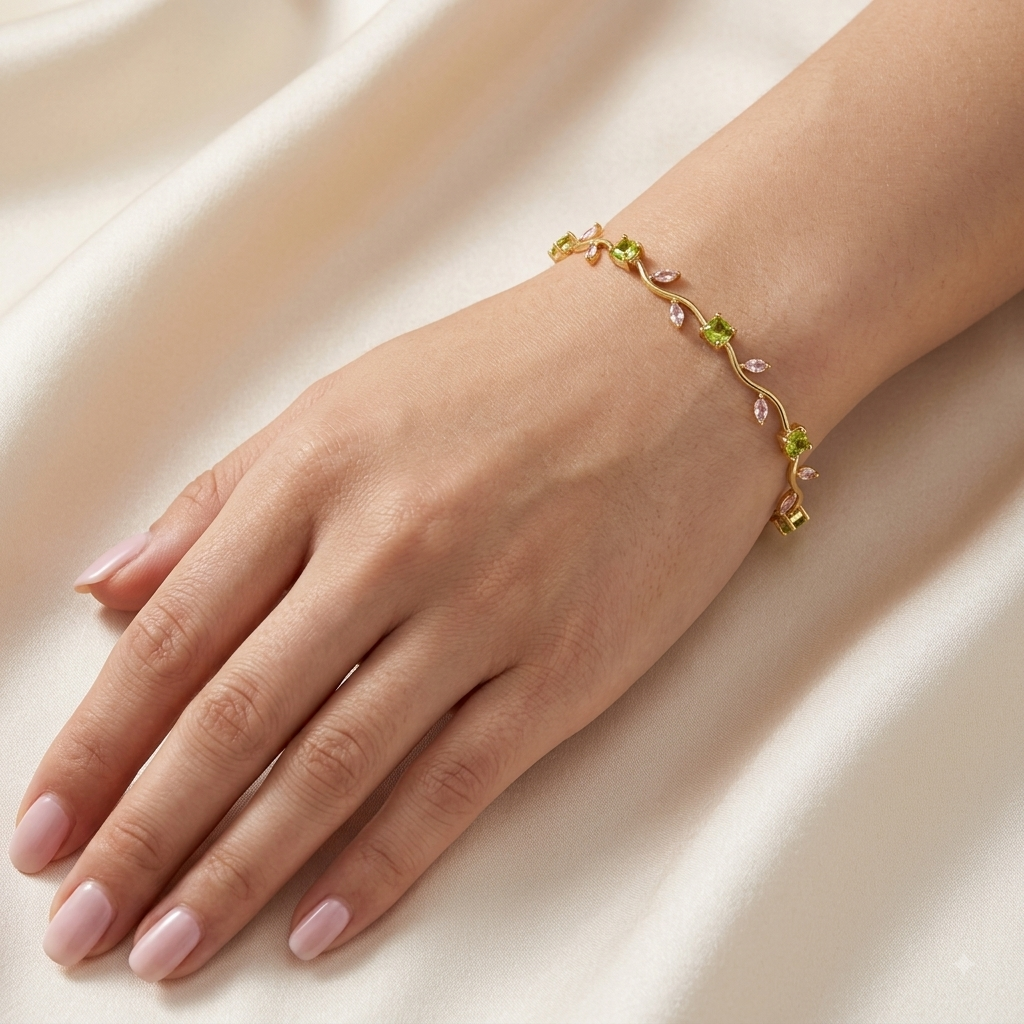 A delicate gold vine bracelet with small green and pink gemstone accents, shown worn on a woman's wrist against a soft cream satin background.