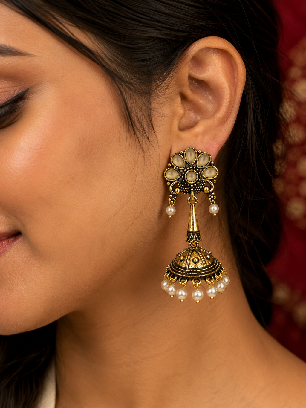 A close-up of a woman's ear wearing an ornate antique gold-toned jhumka earring with cream-colored stones and dangling white pearls, set against a dark hair and red fabric background.