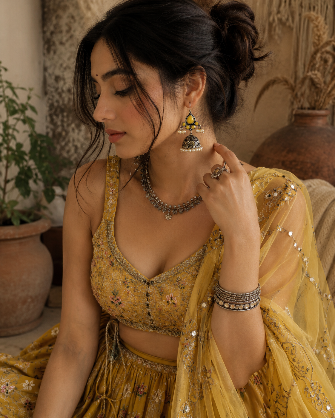 A woman wearing a mustard yellow embroidered traditional outfit with a matching sheer dupatta, styled with silver jewelry including a necklace, earrings, and bangles, in a warm-toned indoor setting.