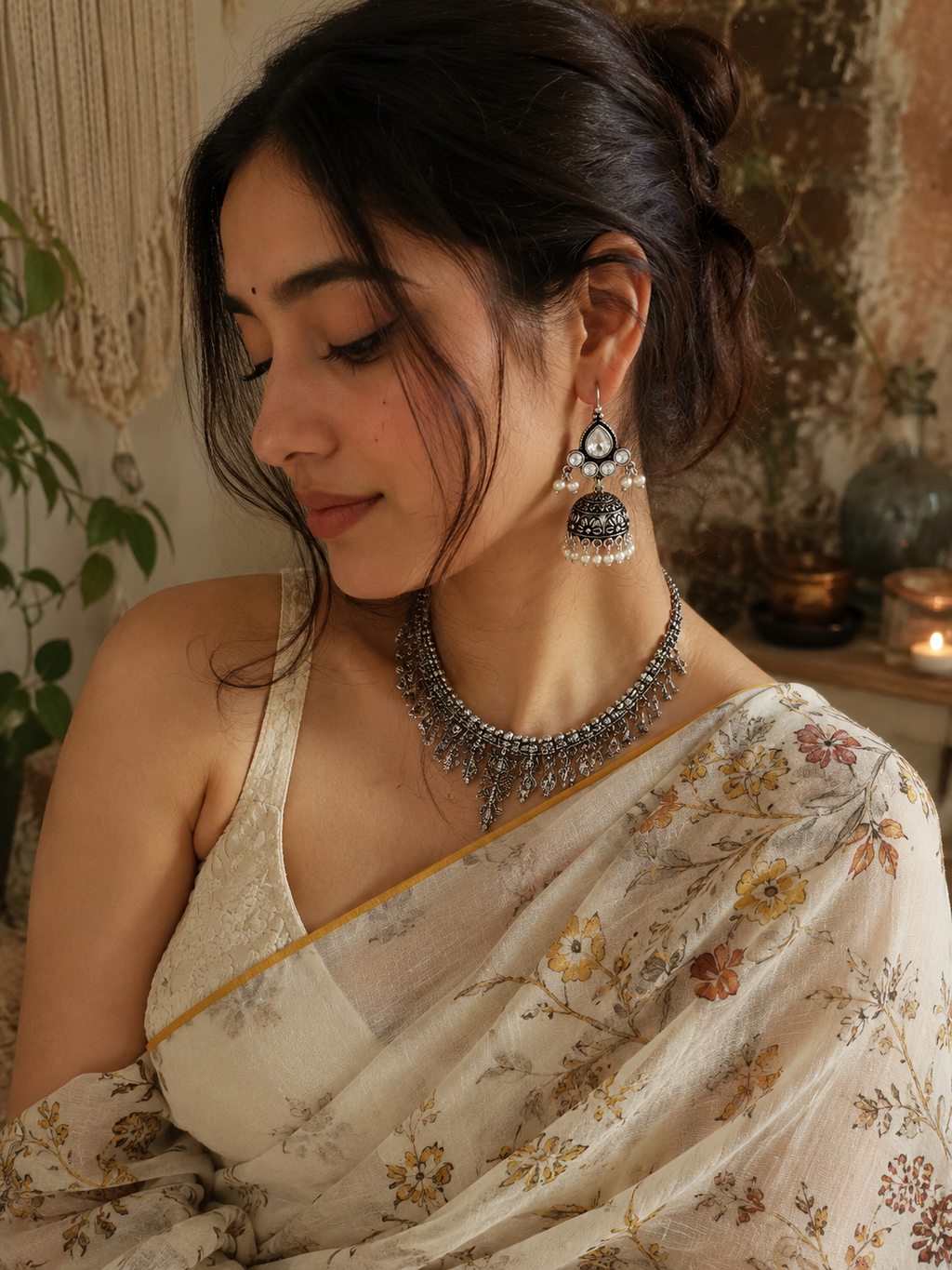 A woman wearing a detailed silver-toned traditional necklace and matching drop earrings, styled with a cream floral saree in a warm, rustic indoor setting.