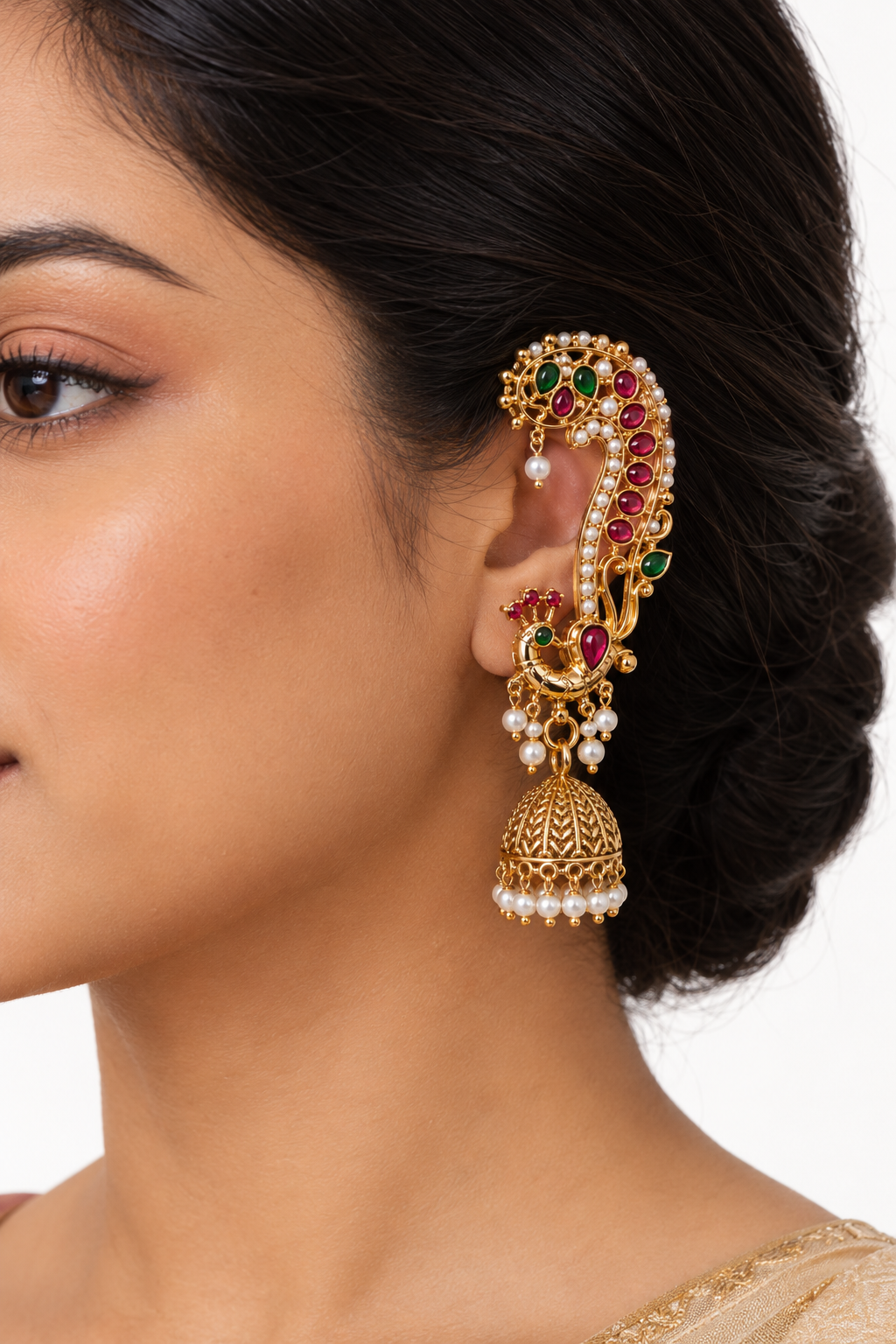 A close-up of a woman's ear wearing an ornate gold-toned traditional Indian ear cuff decorated with red and green stones and white pearl-like beads, with a matching dangling jhumka earring.