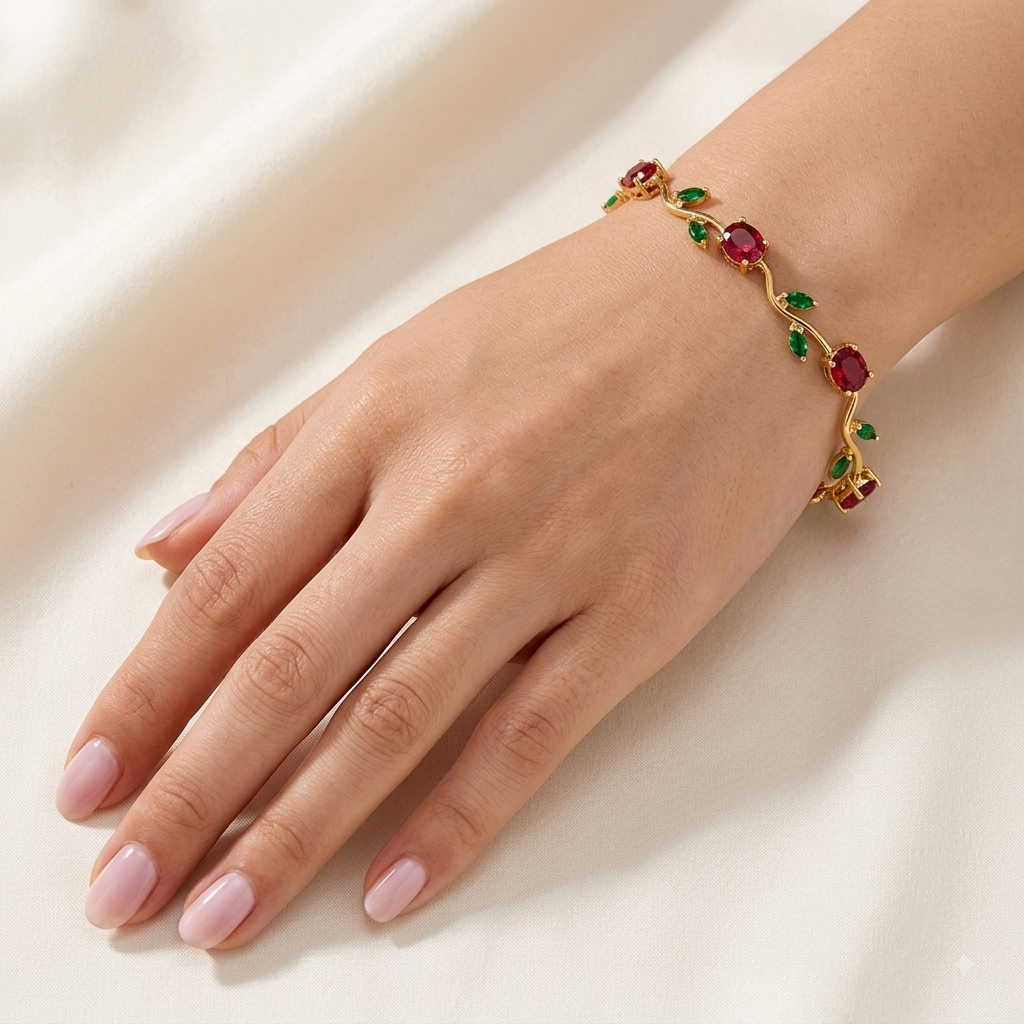 A gold-toned bracelet with oval red gemstones and green leaf accents, shown worn on a woman's wrist with her hand resting beside it against a soft cream fabric background.