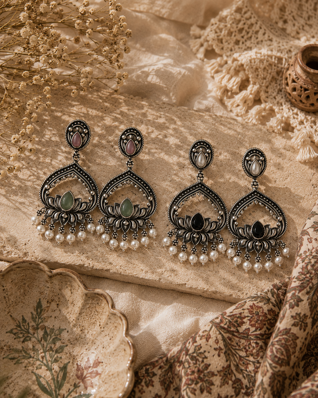 Three pairs of ornate black filigree dangle earrings with green and pink stone accents and white pearl drops, arranged on a beige textured surface with lace, dried flowers, and patterned fabric.