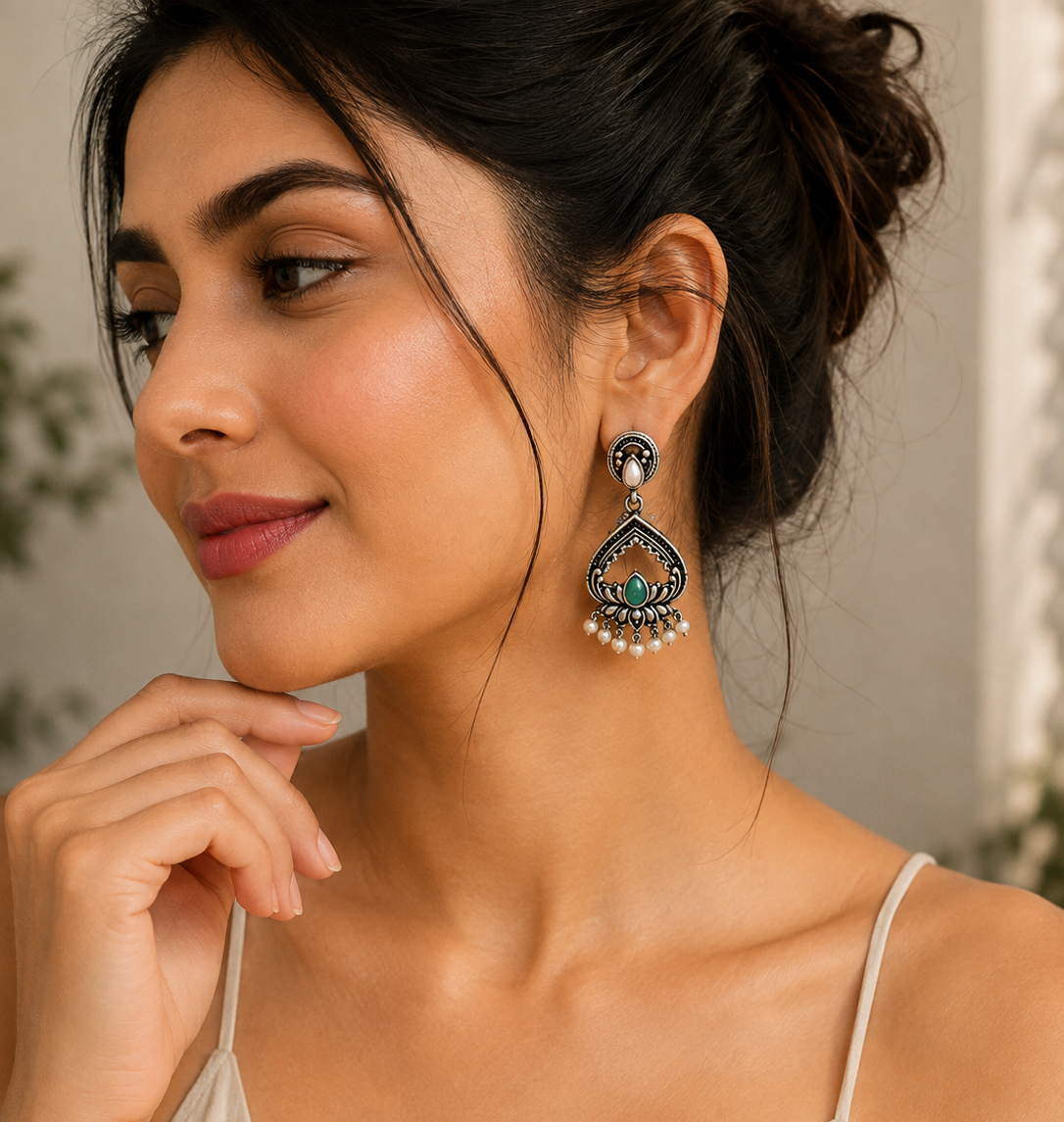 A woman wearing a dangling ornate earring with a silver-toned teardrop design, green stone accents, and small white pearl-like drops, shown in a close-up portrait.