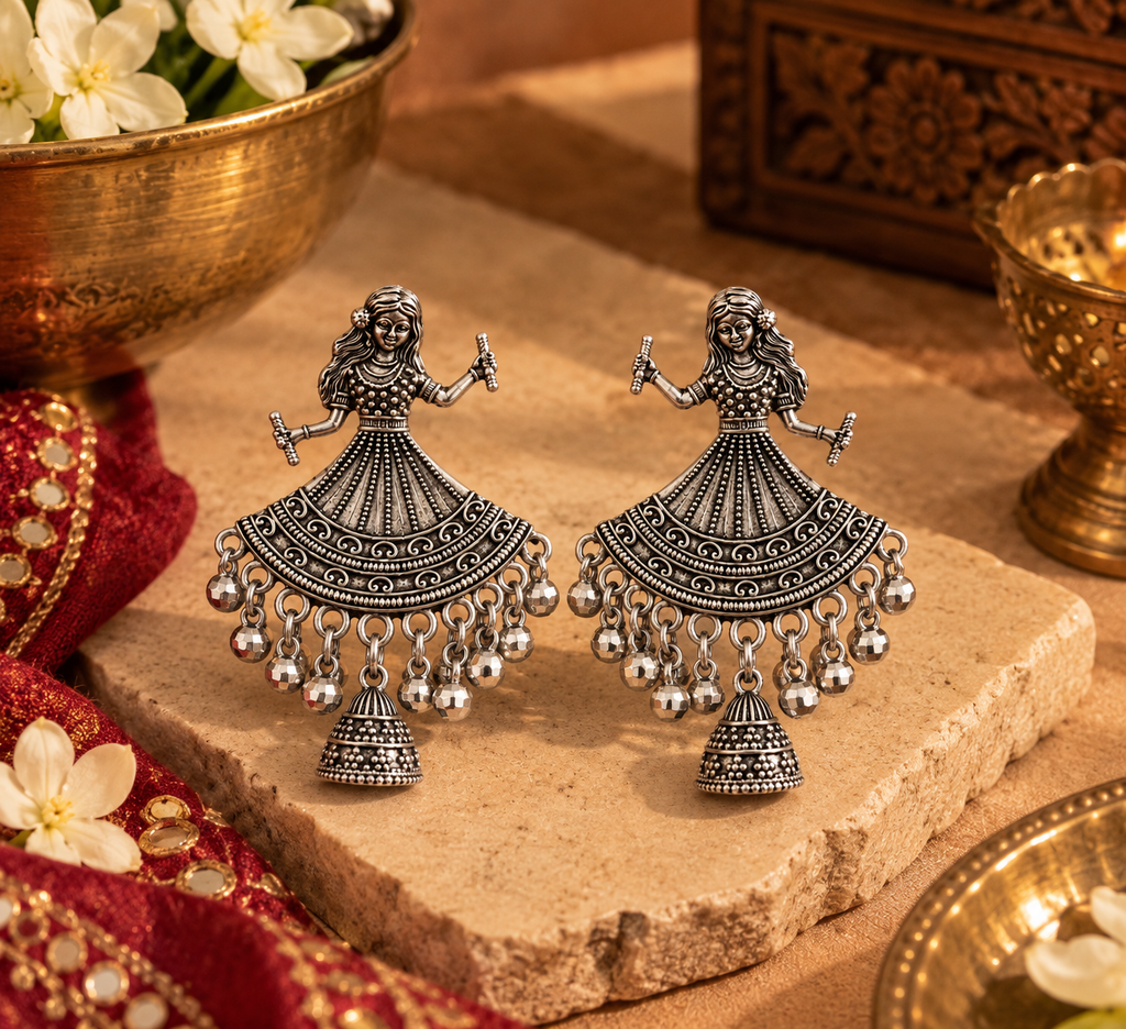 A pair of ornate silver-toned Indian Jhumka earrings with detailed filigree work and dangling bell charms, displayed on a stone slab with traditional decorative props.