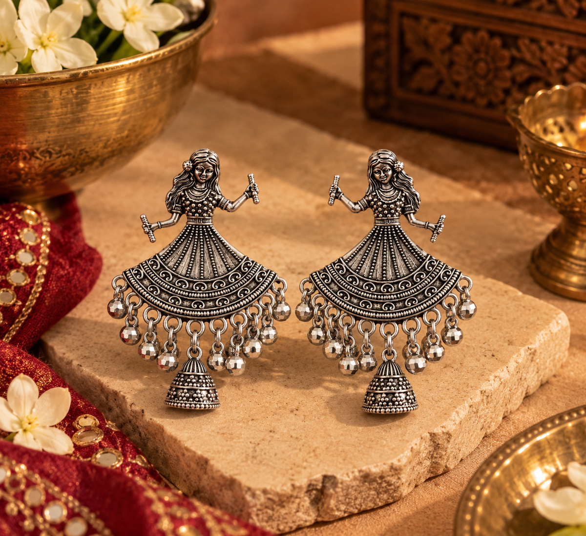 A pair of ornate silver-toned Indian Jhumka earrings with detailed filigree work and dangling bell charms, displayed on a stone slab with traditional decorative props.