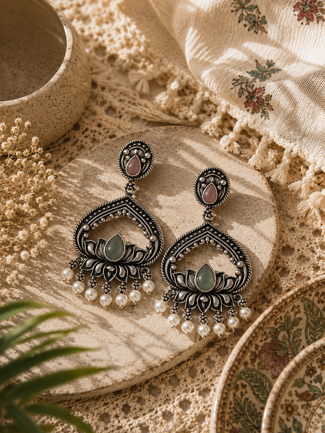 A pair of ornate silver-toned dangle earrings with pink stone accents and green teardrop centers, decorated with dangling pearl beads, displayed on a round wooden tray over a beige lace textile.