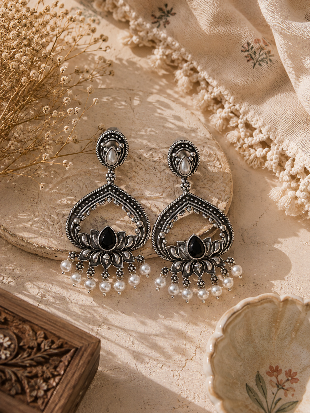 A pair of ornate silver-toned dangle earrings with black inlay, white pearl-like beads, and a central black teardrop stone, displayed on a beige textured surface with dried flowers and fabric.